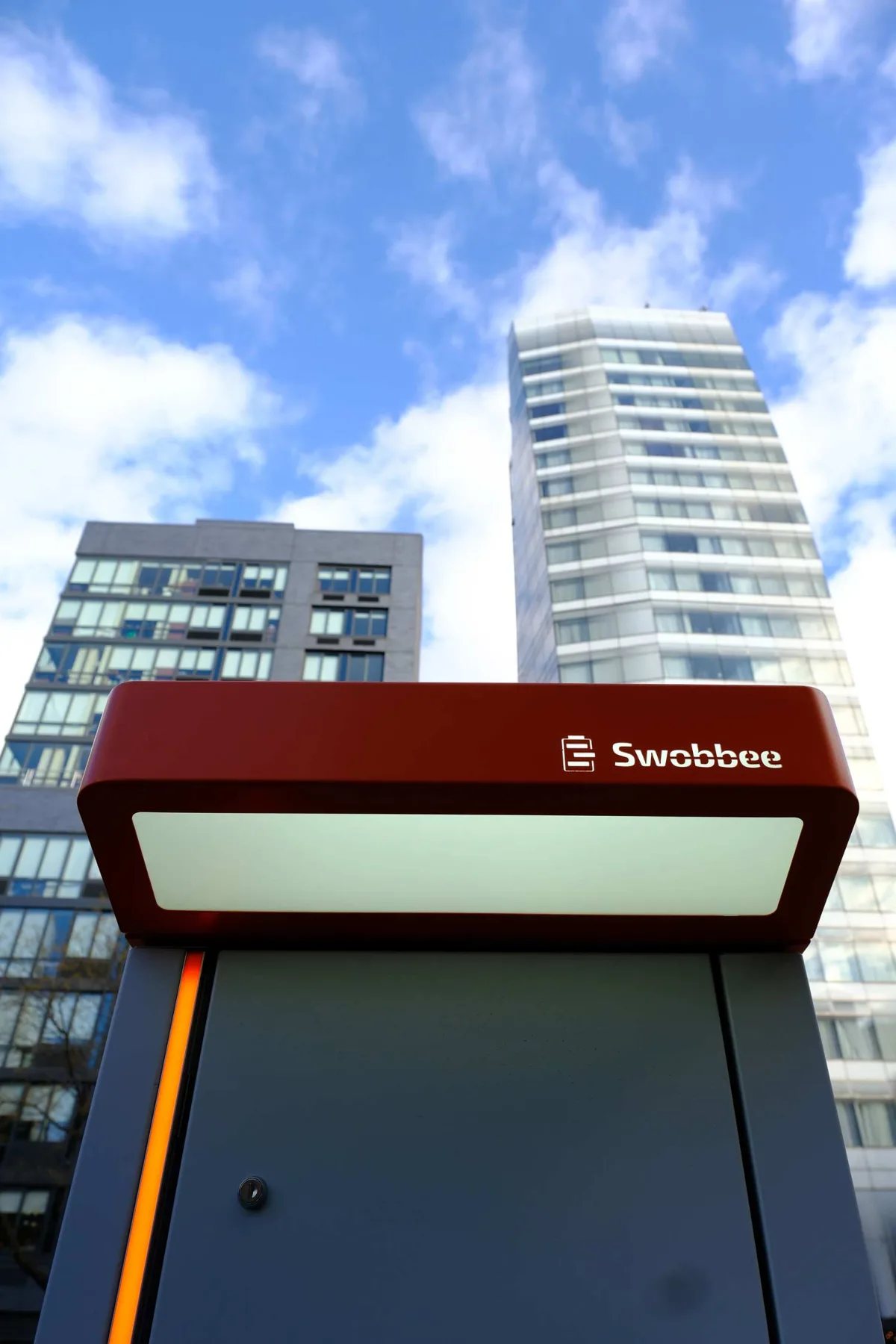 The lit roof of a Swobbee Station with the Swobbee logo, high-rise buildings at Cooper Square, New York, behind it