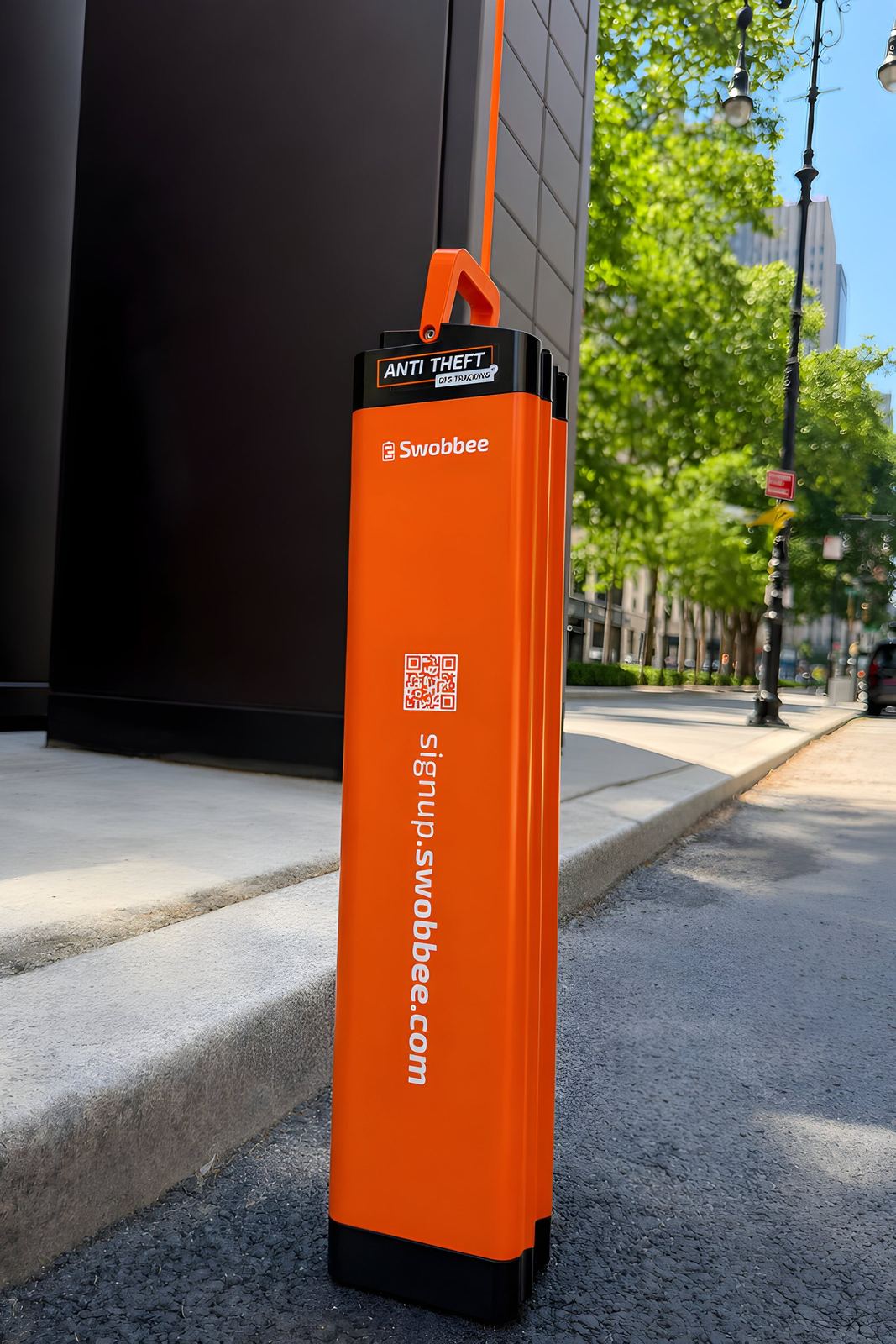 Ein oranger Swobbee One Akku mit Diebstahlschutz-Aufkleber auf dem Gehweg neben einer Station